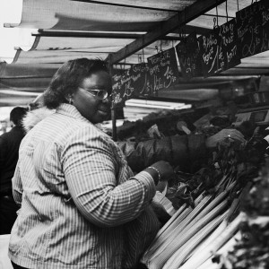 A person wearing glasses and a striped jacket is selecting fresh produce from a market stall at Marché Aligre. The stall has a variety of vegetables, with prices written on chalkboard signs above. The person is focused on choosing items, while other shoppers are visible in the background.