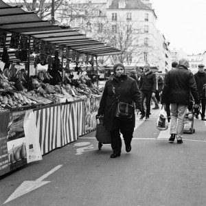 Black-and-white photo of Marché Aligre with several stalls selling produce. A woman wearing a coat and a headscarf walks toward the camera while pulling a shopping cart. Other people are shopping and walking along the market stalls under striped awnings.