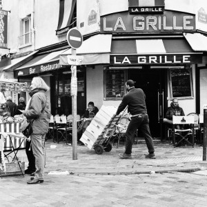 Black and white photo of a street scene outside "La Grille" restaurant near Marché Aligre. A person pushes a hand truck loaded with boxes while another stands nearby, and several people sit at outdoor tables. A street sign and storefronts are visible in the background.