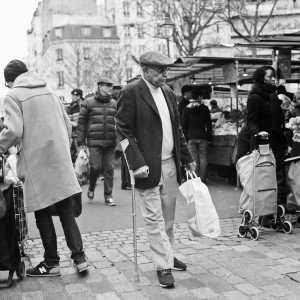 A black-and-white photo of a bustling market scene at Marché Aligre. A man with a crutch carries a grocery bag while walking through the cobblestone street. Around him, people in winter clothing pull shopping carts. Buildings and market stalls form the backdrop.