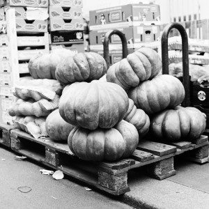 A stack of large pumpkins displayed on a wooden pallet at Marché Aligre. Boxes of produce are visible in the background. The setting appears to be outdoors, and the image is in black and white, capturing the timeless charm of this Paris market.