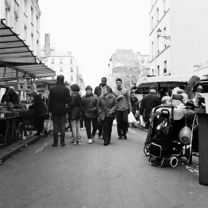 A bustling Marché Aligre street market scene in black and white. People are walking and browsing various stalls and stands along the side of the street. Some are carrying shopping bags, and a stroller is visible to the right. Urban buildings line both sides of this vibrant Paris market.