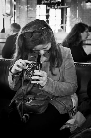 A woman sits indoors on a bench, looking down as she adjusts a vintage twin-lens camera. Wearing a jacket with sunglasses on her head, she appears focused—capturing the essence of an Alix Photographe moment amid blurred figures in the background.