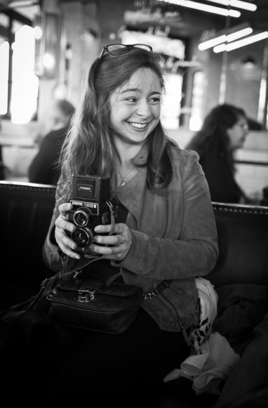 A smiling woman sits indoors on a couch, holding a vintage twin-lens reflex camera. She has long hair, wears a jacket, and glasses rest on her head. In the blurred background, people and lights surround Alix Photographe at work.