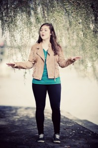 A woman stands outdoors on a stone path beneath hanging branches, wearing a tan jacket, teal shirt, black pants, and sneakers, with her arms outstretched and mouth open as if speaking or singing.