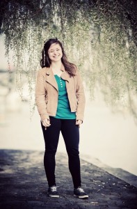 A young woman stands outdoors on a stone path, smiling under hanging green branches. She wears a tan jacket, green shirt, black pants, and black shoes with white laces. The background is softly blurred.