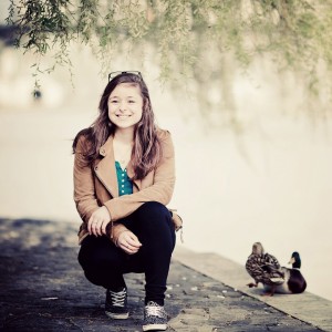 A young woman with long brown hair, wearing a brown jacket and black pants, crouches on a riverside path and smiles at the camera. Two ducks are standing nearby, and leafy branches hang above her.