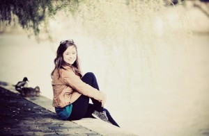 A young woman sits on a stone ledge by the water, smiling at the camera. She is wearing a brown jacket, black pants, and patterned shoes. There are ducks nearby and overhanging tree branches above her.