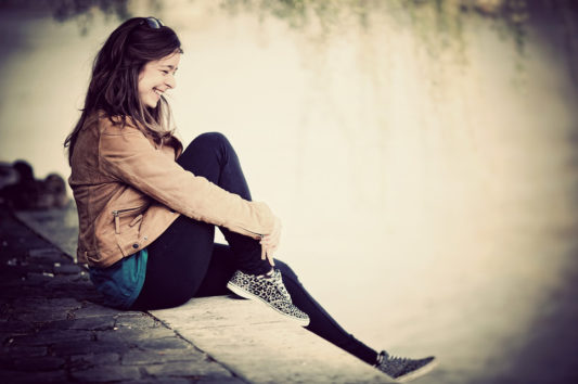 A young woman sits on a stone ledge by the water, smiling and looking relaxed. She wears a brown jacket, black leggings, and leopard-print sneakers, with her long hair down and sunglasses on her head.