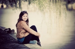 A young woman sits on a stone ledge by the water, smiling at the camera. She wears a brown jacket, black pants, and blue sneakers. Sunglasses rest on her head, and trees hang over the water in the background.