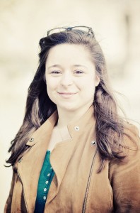 A young woman with long brown hair, wearing a tan suede jacket over a green top, stands outdoors and smiles softly at the camera. Glasses rest on her head. The background is bright and blurred.