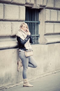 Joanna Mirven, in a scarf, leather jacket, and boots, leans against a stone wall, talking on her phone. She stands casually with one leg bent, next to a barred window on a city street.