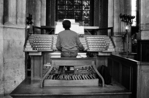 An organiste sits at the large pipe organ in Saint-Eustache, facing away from the camera, playing music in a grand cathedral. The organ has numerous keys, stops, and foot pedals. The surrounding setting includes stone walls and a stained glass window in the background.