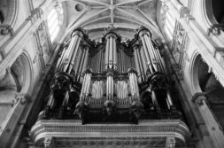 Black and white photo of a magnificent pipe organ inside the grand Saint-Eustache cathedral. The view is from below, emphasizing the intricate details of the pipes and the ornate architecture surrounding the organ. Tall windows and vaulted ceilings add to the grandeur, awaiting an organist's touch.