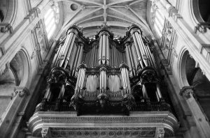 Black and white photo of a magnificent pipe organ inside the grand Saint-Eustache cathedral. The view is from below, emphasizing the intricate details of the pipes and the ornate architecture surrounding the organ. Tall windows and vaulted ceilings add to the grandeur, awaiting an organist's touch.
