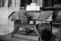 An organist is sitting and playing a large pipe organ in what appears to be a church or cathedral. The organ has multiple rows of keys, numerous stop knobs, and several foot pedals. Music sheets are open on the music stand in front of the player, resembling a scene from Saint-Eustache Cathedral.