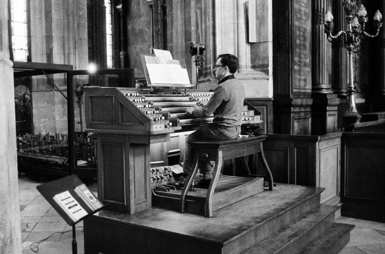 Black and white photo of an organiste playing a large pipe organ in Saint-Eustache Cathedral. The person is seated on a bench with their hands on the organ's keyboards and their feet on the pedals. Tall columns and ornate details of the cathedral surround them.