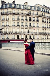 A couple stands close together on a city street; the mariée wears a long robe rouge and the man wears a dark suit. Behind them is an ornate, light-colored European-style building with many windows and balconies.