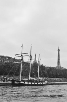 A black and white photo of the trois-mâts La Boudeuse docked along the river, with trees in the background and the Eiffel Tower visible in the distance under a cloudy sky.