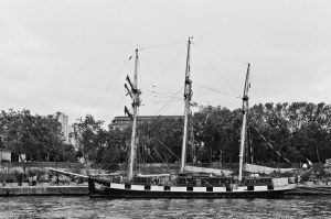 A black and white photo of the three-masted sailing ship La Boudeuse docked along a waterfront, with trees and buildings visible in the background.