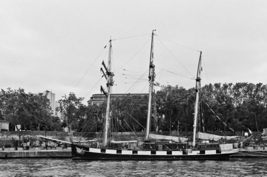 A black and white photo of the three-masted sailing ship La Boudeuse docked along a waterfront, with trees and buildings visible in the background.