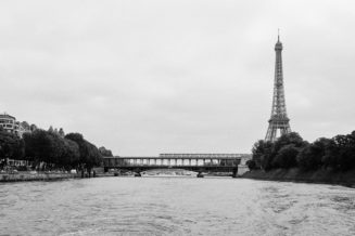 Black and white photograph of the Tour Eiffel in Paris, France, as seen from the Seine River. The tower stands tall on the right side, with a bridge featuring an RER train passing in the foreground and tree-lined riverbanks leading the view towards the background.