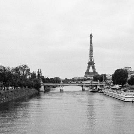 A black and white photograph of the Tour Eiffel in Paris, France. The tower stands tall in the background, with a river and a bridge in the foreground. Boats are docked along the riverbank, and trees line both sides of the river, while an RER train crosses the bridge.