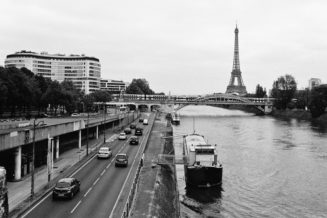 A black and white photo of the Seine River in Paris, with the Tour Eiffel in the background. Cars are driving on the road by the river, and there is a boat moored in the river. A bridge crosses near the Tour Eiffel, while buildings and trees line the road, capturing a moment of harmony beside an RER route.
