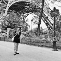 A black-and-white photo of a person standing in front of the Eiffel Tower. The individual is engrossed in reading or looking at something in their hands, standing by a pathway with a sign that reads "Avenue Gustave Eiffel," surrounded by trees and greenery.