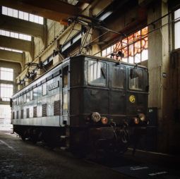 A black vintage train engine is displayed indoors on train tracks at the dépôt de la Chapelle. The engine has a lit headlight and various mechanical details. The industrial space, part of the eclectic Ground Control exposition, features large windows that allow natural light to illuminate the train.
