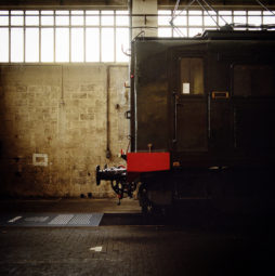 A vintage, dark-colored train car rests inside the industrial ambiance of dépôt de la Chapelle. The car's coupling mechanism faces a textured, worn concrete wall. Overhead windows let in soft light, illuminating the scene. The floor is paved with aged bricks, contributing to the timeless atmosphere of this Ground Control Exposition.