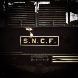 Close-up of the front of a black train with a sign reading "S.N.C.F." The background showcases the train's grilles and reflective panels, indicating it is stationary and possibly displayed indoors, perhaps at an Exposition Ground Control or dépôt de la Chapelle, given the lighting.