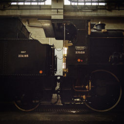 A close-up of a black steam locomotive with identification numbers "230-09" and "23A165" on the side, partially visible in the dimly lit environment of dépôt de la Chapelle, showcasing the intricate details of its mechanical components and structure.