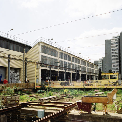 An urban rail yard scene with several sets of tracks converging near a large, industrial building reminiscent of the dépôt de la Chapelle. People are seen walking near the building, which has multiple levels and large windows. High-rise buildings are visible in the background under a partly cloudy sky.
