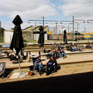 People relax on lounge chairs beneath large umbrellas beside train tracks on a sunny day. The background features multiple train lines, including some with stationary trains near dépôt de la Chapelle, and an industrial setting with wires and buildings. The sky is partly cloudy.