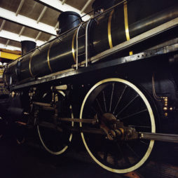 A close-up view of the side of an antique steam locomotive, showcasing its large, black wheels with white rims and metallic components. The locomotive, part of the Exposition at Ground Control's dépôt de la Chapelle, is housed in a well-lit museum with a high, white ceiling.