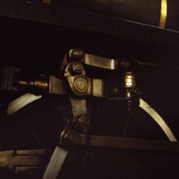 Close-up of the intricate metal components of a vintage steam locomotive's wheel assembly at the dépôt de la Chapelle, showing gears, bolts, and levers with dark, moody lighting. The mechanical parts are highlighted, emphasizing their complexity and craftsmanship.