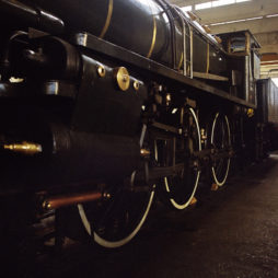 A close-up view of the side of a vintage black steam locomotive at dépôt de la Chapelle, showcasing its large wheels with white rims. The locomotive is positioned indoors, with sunlight streaming in through the windows above, casting shadows on the metal surface.