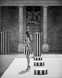 A woman in a striped robe Buren and heels stands among tall, black-and-white striped columns in an outdoor plaza with a stone building in the background.