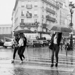 Black and white photo of three people walking on a rainy Paris street; two share an umbrella and laugh, while one walks alone holding an umbrella. Cars and buildings fade into the misty pluie Paris background.