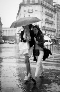 Two women walk closely together under an umbrella on a rainy Paris street, carrying shopping bags. The wet pavement glistens as buildings and vehicles line the background. The black and white scene captures the charme de la pluie in the city.