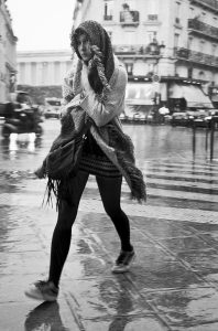 A woman walks briskly across a wet city street under the pluie à Paris, covering her head with a scarf and wearing a patterned skirt, leggings, sneakers, and a bag, with blurred buildings and cars in the background.