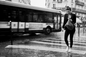 A woman walks quickly across a wet street in heavy pluie, clutching her bag, as a city bus passes by in the blurred urban backdrop of Paris.