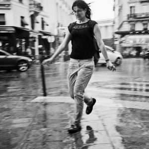 A woman with wet hair and clothes briskly walks through a rainy Paris street. Her long stride cuts through the pluie-soaked scene, while blurred buildings and cars in the background emphasize her motion and the damp atmosphere.