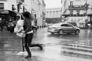 A woman runs across a wet Paris street in the rain, carrying bags. Cars pass by as the pluie tombe, and buildings and shops line the background. The photo is black and white, capturing a sense of motion and urgency.