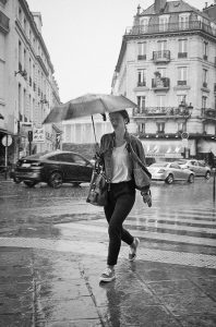 A woman walks quickly across a city street in the rain, holding an umbrella—classic pluie Paris. Cars drive by in the background, and the wet sidewalks glisten in this black-and-white scene.