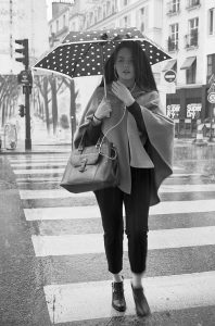 A woman walks across a crosswalk in the pluie, holding a polka-dot umbrella and a handbag. She wears a cape-style coat and looks forward, with Parisian buildings and shops visible in the background.