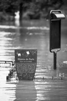 A historic sign and a mailbox are partially submerged by crue floodwater in Paris. The surrounding area, including a fence at Square du Vert-Galant, is heavily flooded. The black and white scene highlights the severity of the inundation.