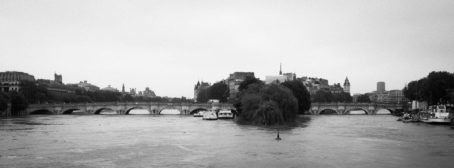 A black and white photo of the Seine River in Paris, featuring boats and historic buildings. The Square du Vert-Galant is visible near a multi-arched bridge, all beneath an overcast sky reflecting the mood after a recent crue.