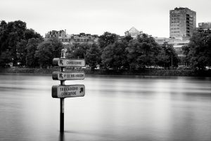 Black and white photo of street signs, including Direction Tour Eiffel, partly submerged in water, pointing to Cambronne, Pt de Bir-Hakeim, Trocadéro, and Concorde, with trees and urban buildings in the background.